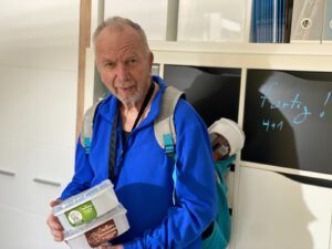 Ein Mann in blauer Jacke steht für einem hellen Bücherregal. Er hält Eisdosen in der Hand, aus seinem Rucksack schaut eine weitere heraus.