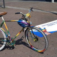 Das Bild zeigt ein Fahrrad, dass in Regenbogenfarben besprüht ist, neben einem großen, auf dem Boden liegendem Schriftzug anlässlich des Internationaler Gedenktags gegen Homo- und Transphobie (IDAHOBIT).