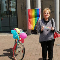 Frau Funke steht neben einem bunten Fahrrad, an welchem Luftballons hängen. Im Hintergrund ist eine Regenbogenfahne anlässlich des Internationaler Gedenktags gegen Homo- und Transphobie (IDAHOBIT) zu sehen.