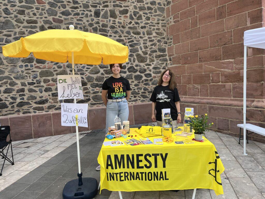 Gelber Stand von Amnesty International mit 2 Frauen dahinter.