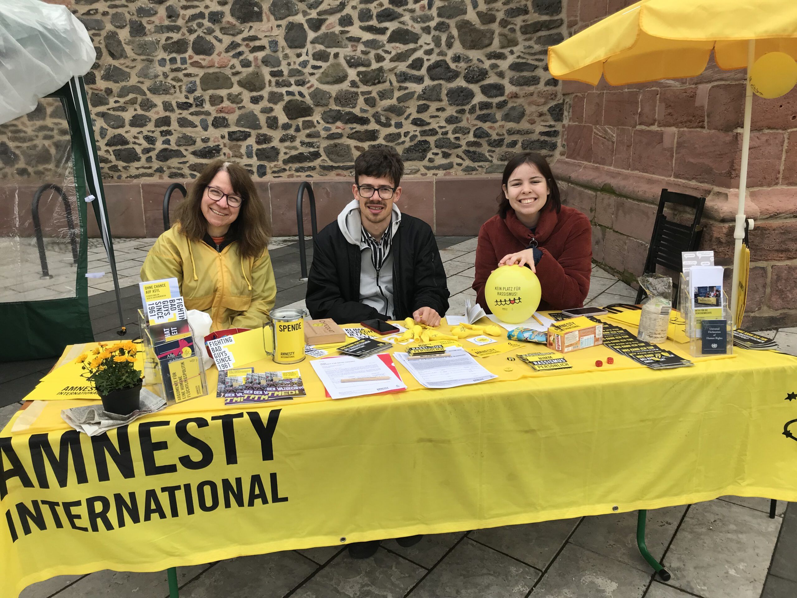Tisch mit gelben Decktuch auf dem Amnesty International steht mit 3 Personen die dahinter sitzen