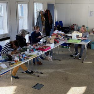 Vier Frauen sitzen an einem langen Tisch und malen, stricken, nähen.