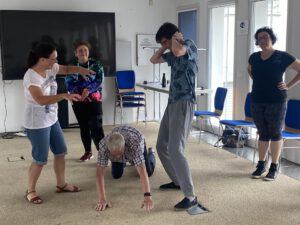 2023: Improvisationstheater-Workshop. Drei Frauen und zwei Männer stellen etwas dar. Einer der Männer ist auf dem Boden im Vierfüßler-Stand, einer steht daneben und hält sich die Ohren zu. Die Frauen lachen.