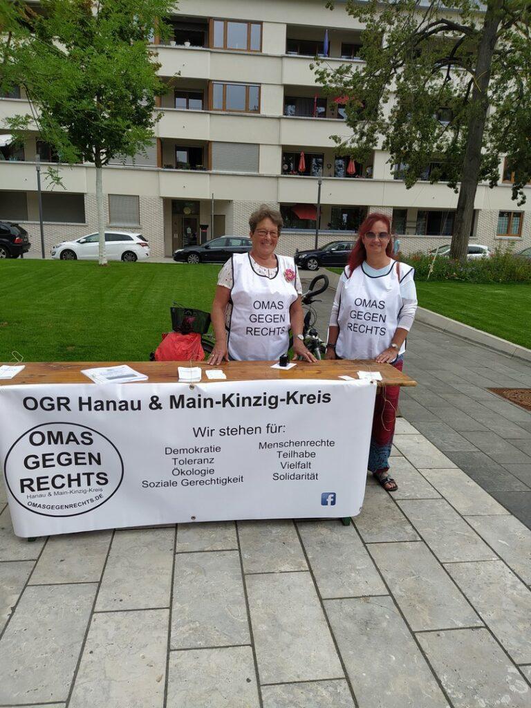 Zwei Omas gegen Rechts stehen in ihrem weißen T-Shirt hinter einem Tisch. Am Tisch hängt ein Banner mit Werbung für die Omas gegen Rechts (OGR) Hanaus und des Main-Kinzig-Kreises.