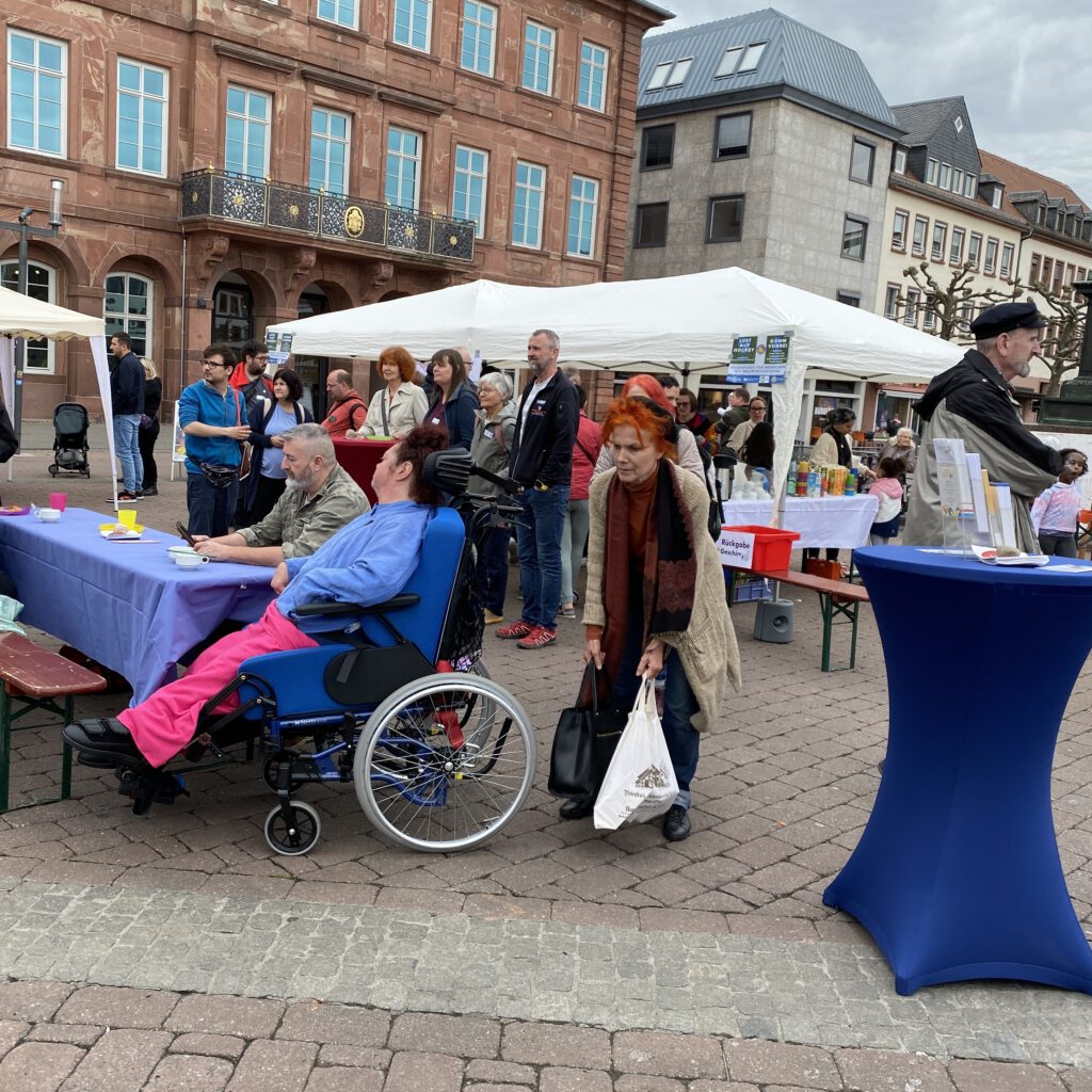 Im Rahmen des jährlichen Europäischen Protesttags zur Gleichstellung von Menschen mit Behinderungen, "Dinner in bunt" 2024. Vor dem Bürgerhaus am Marktplatz versammelte sich eine große Menschenmenge vor Infoständen, Essen- und Getränkeständen sowie sitzend auf Sitzbänken.