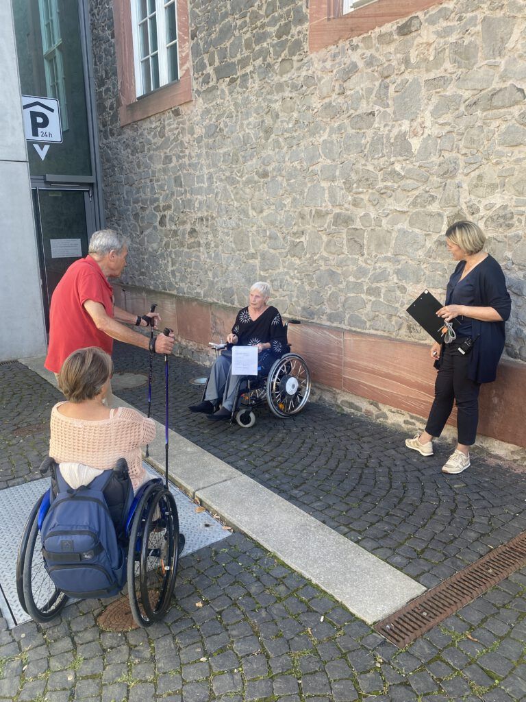 2 Frauen im Rollstuhl, ein älterer Herr mit Stöcken und eine Frau mit Klembrett schauen sich den Extra-Zugang an der auf Kopfsteinpflaster ins Gebäude führt.