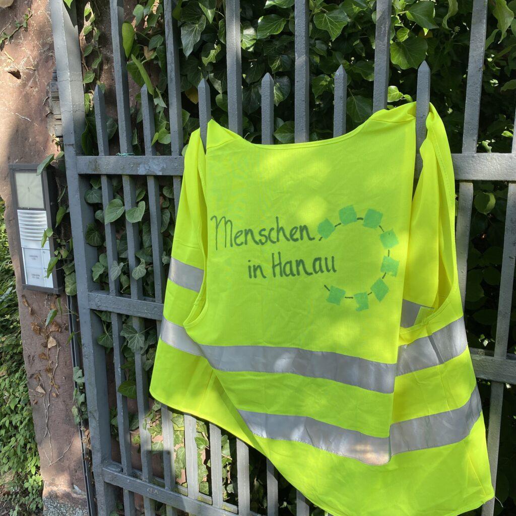 Eine gelbe Warnweste mit der Aufschrift "Menschen in Hanau" hängt an einem Zaun.