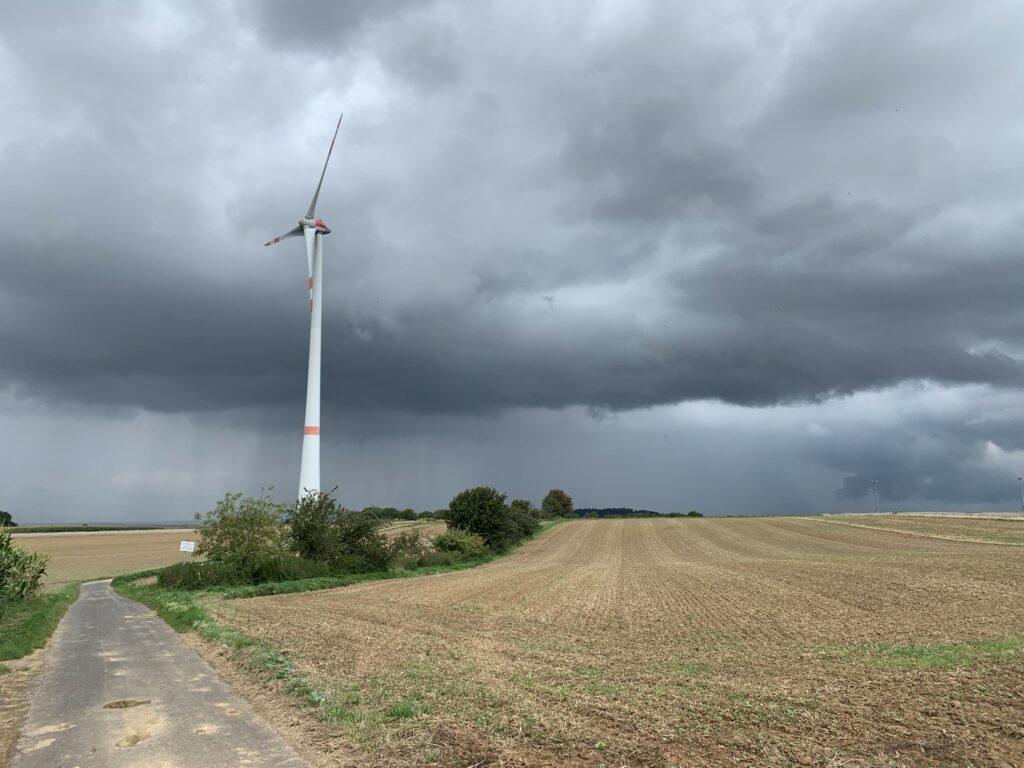 Das Bild zeigt ein Windkraftrad und dahinter sehr dunkle Wolken mit kräftigen Regenschauern.
