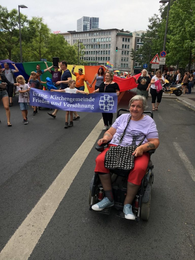 Das Bild zeigt eine Teilnehmerin, die mit der Parade mit läuft. Sie wird bald 70 Jahre und sitzt im Rollstuhl.