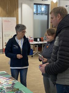 Iris im Gespräch mit einer Frau und einem Mann. Iris trägt eine blaue Jacke mit der Aufschrift Menschen in Hanau.