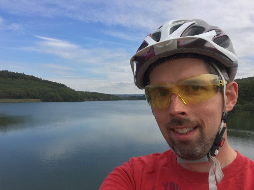 Daniel mit Fahrradhelm und schützender Brille vor Insekten auf dem Staudamm der Kinzigtal-Sperre. Im Hintergrund sieht man das aufgestaute Wasser.