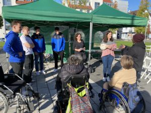 2023: Mehrere männliche Jugendliche der HSG Handballabteilung hören und sehen dem MiH-Team zu. Eine gehörlose Frau und eine Gebärdendolmetscherin sprechen miteinander. Zwei Frauen im Rollstuhl schauen zu. Das Bild entstand auf dem Tauschmarkt vor der Wallonisch-Niederländischen Gemeinde.