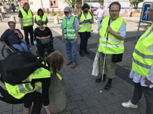 2020: Freiwilligentag. Mehrere Personen mit Warnwesten stehen um einen Poller herum. Ein Mann hat einen Taststock. Eine Frau klebt Kontraststreifen auf einen Poller.