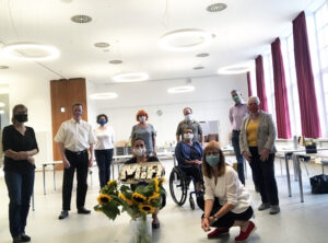28. August 2020: Gründungssitzung von Menschen in Hanau. Neun Personen sind in einem Raum und tragen Corona-Masken. Eine der Personen, eine Frau, sitzt im Rollstuhl. In der Mitte von allen steht ein Strauß Sonnenblumen.