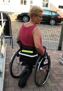 SmartDrive ist unten als schwarzes Rad zu erkennen. Das Bild zeigt den Rollstuhl von hinten. Rita sitzt in ihm.
