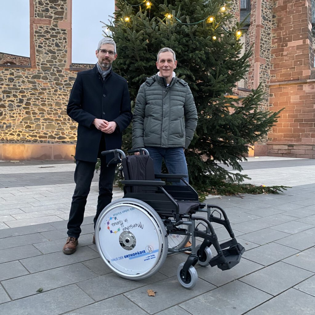 2 Männer stehen zusammen vor einem Weihnachtsbaum und davor steht der gespendete Rollstuhl