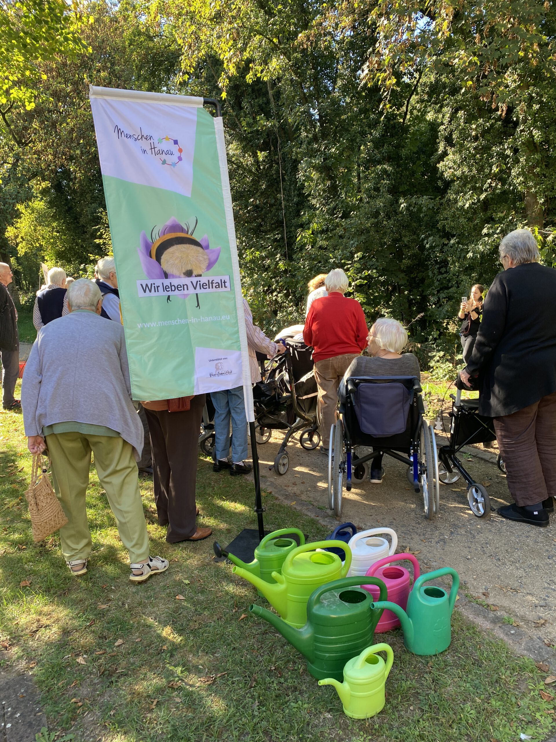 Ein Banner des Vereins mit Hummel-Motiv steht auf der Wiese, davor mehrere bunte Gießkannen, dahinter Bewohner:innen.