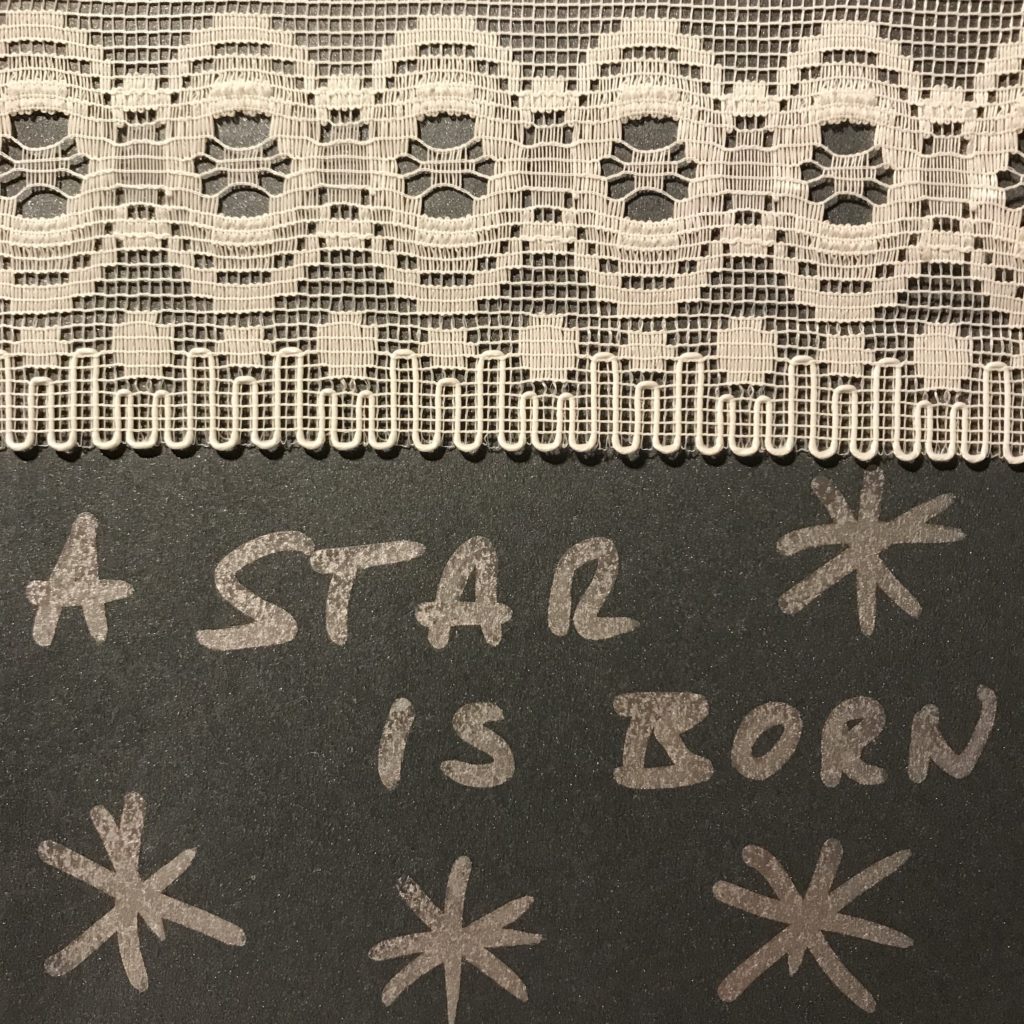 Eine silberne Postkarte auf der steht " A star is born" oben am Rand ist eine weiße Spitze.