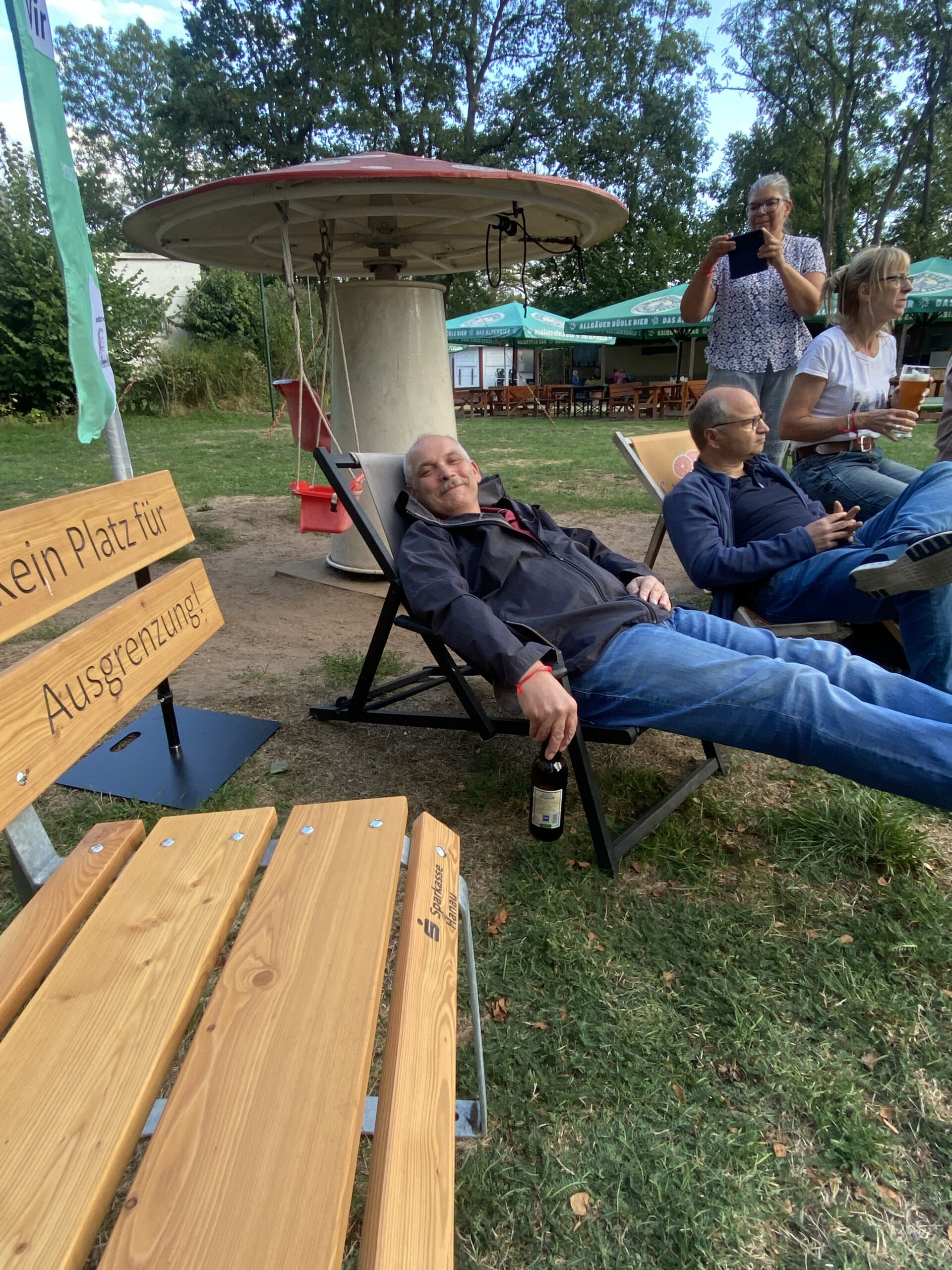 Blick über die Holzbank „Nimm Platz für Vielfalt“ auf Gäste, die in Liegestühlen auf der Wiese entspannen.