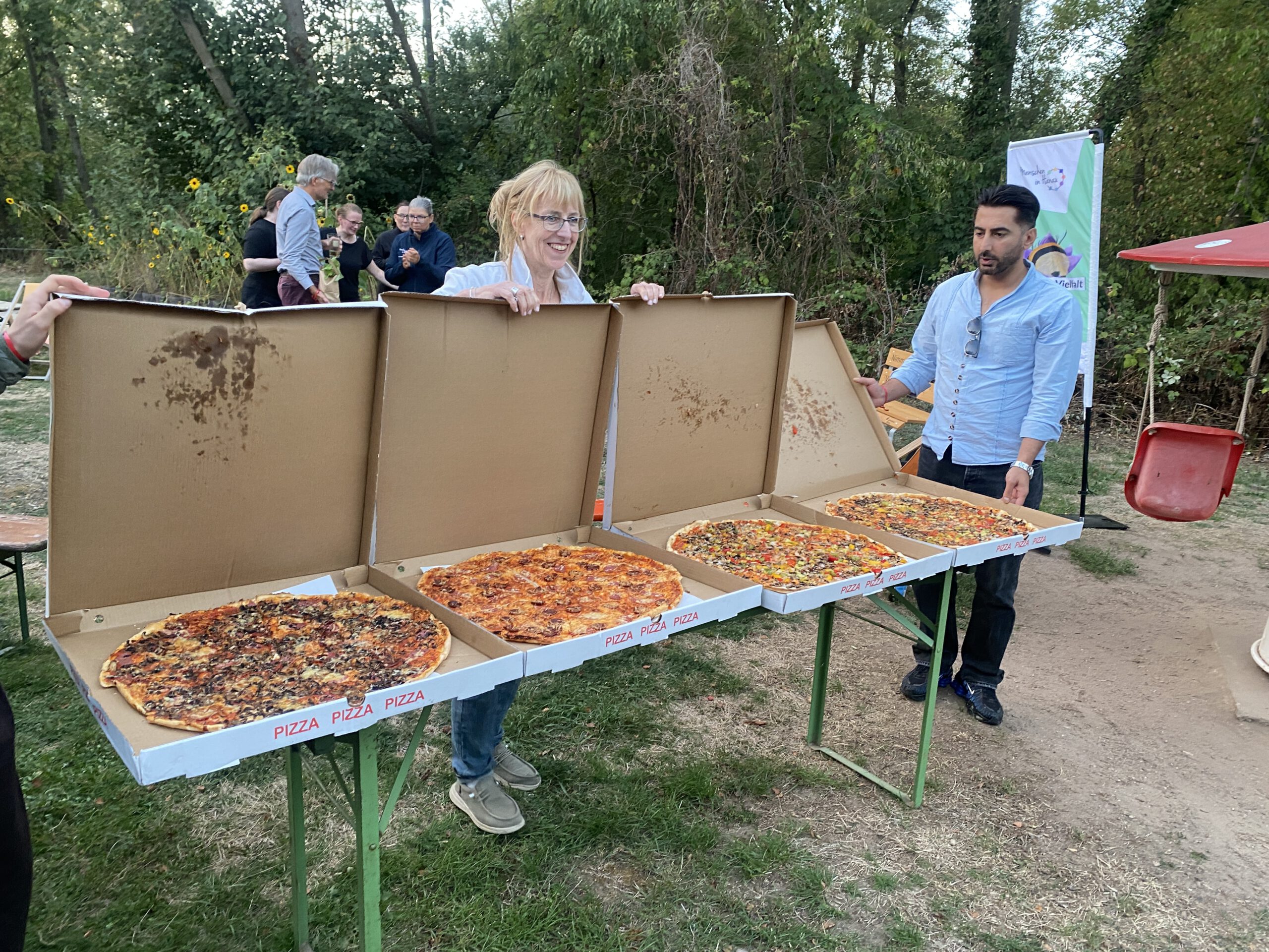 Auf einem langen Tisch liegen mehrere große Pizzen in geöffneten Kartons, daneben stehen Gäste.