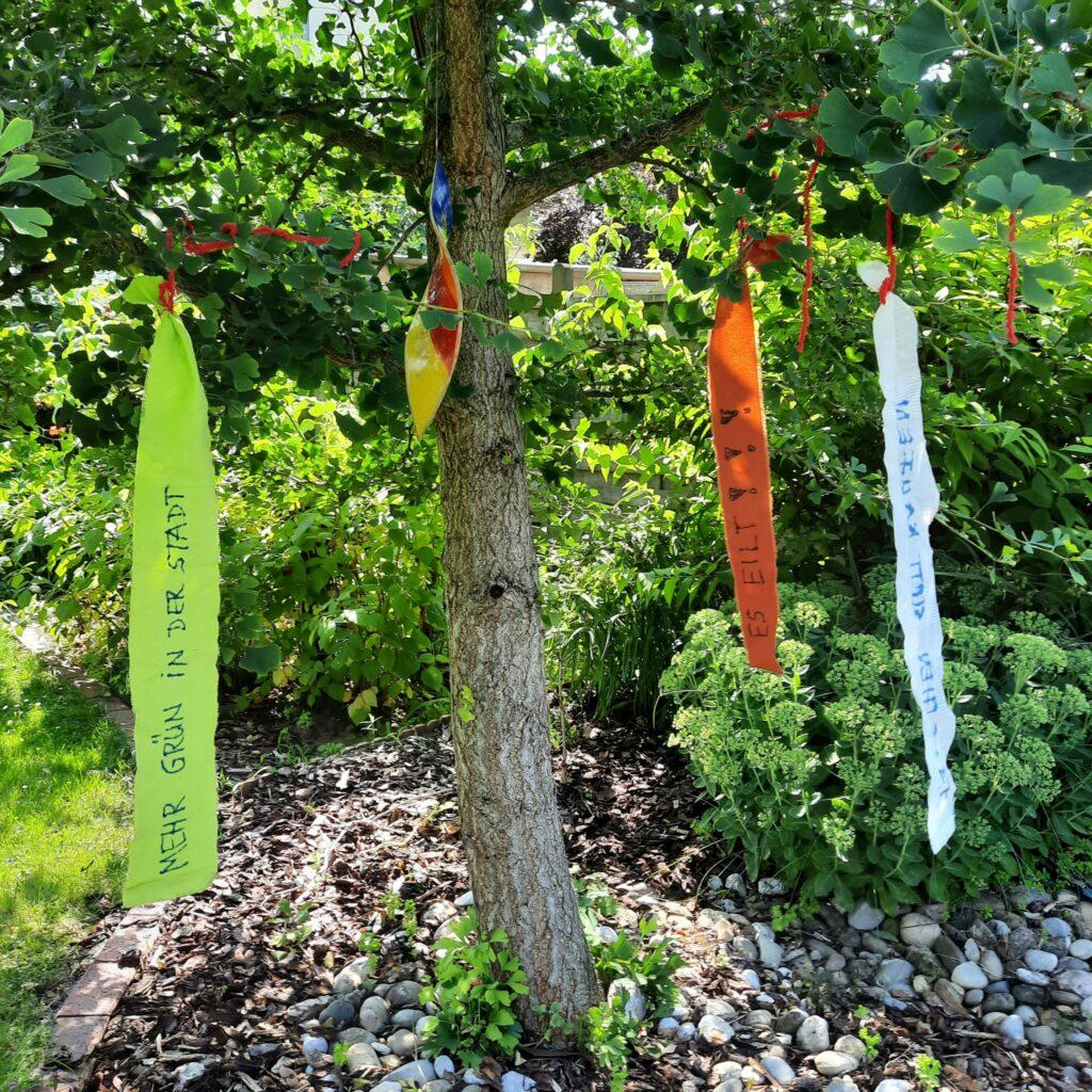 An einem Baum flattern Klimabänder, auf denen Wünsche zum Klima stehen.