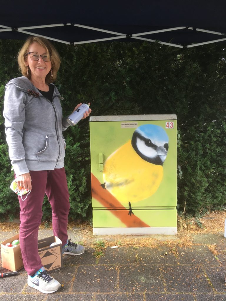 Mit einer Spraydose bewaffnet steht diese Dame neben einem Stromkasten, auf dem ein gelber Vogel mit blauem Köpfchen auf einem Ast sitzt.
