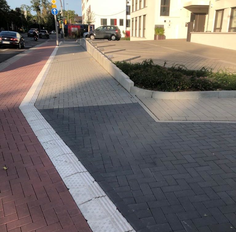 Zu sehen ist ein roter Fußgängerweg, der durch einen weißen Streifen mit Rillen von dem grauen Fußweb getrennt ist.