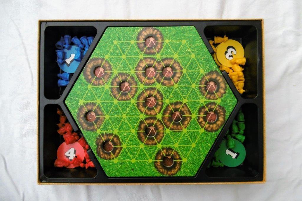 Spielbrett im Karton. Vier Farben sind am Rand als Figuren ausgeführt. In der Mitte auf dem Spielbrett sieht man Maulwurfhügel mit einem Loch in der Mitte.