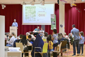 2025: Bingo. Viele Menschen sitzen im Kulturforum an Tischen. Ein Moderator steht auf einer Bühne. Hinter ihm eine Leinwand, auf der Bingo-Zahlen gezeigt werden.