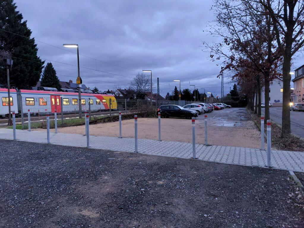 Eiin Fußweg ist mit kontrastreiche gestalteten Pollern von den umgebenden Parkplätzen geschützt.. Im Hintergund ist eine Regionalbahnam am Bahnsteig zu erkennen.