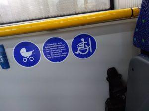 Wir sehen eine gelbe Haltestange im Mehrzweckbereich eines Buses. Unter der Stange sind 3 Piktogramme angebracht, die darauf hinweisen, dass die Fläche für Kinderwagen, Rollstuhlfahrer und Rollatoren freizuhalten ist.