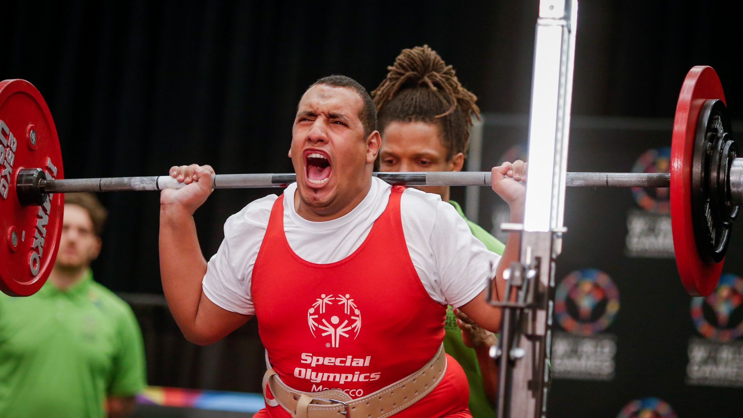 Mann der beim Gewichteheben aufschreit vor Anstrengung. Trägt ein rotes T-Shirt mit den Special Olympics daraufstehend.