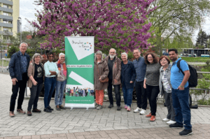 Eine Gruppe von Menschen steht aufgereiht neben einem grünen Menschen in Hanau Banner. Im Hintergrund ist ein Baum der lilane Blüten trägt. 