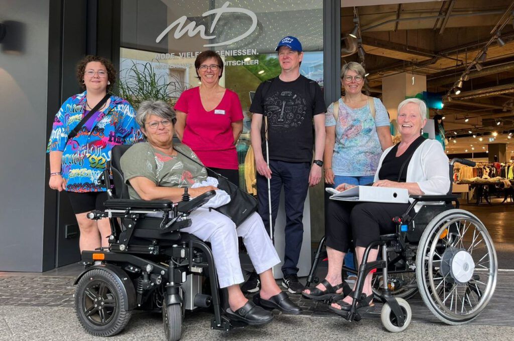 Eine Gruppe steht zum Gruppenfoto vor dem Müller Dietschler in Hanau. 2 Damen sitzen im Rollstuhl und ein junger Mann trägt einen Blindenstock mit sich.