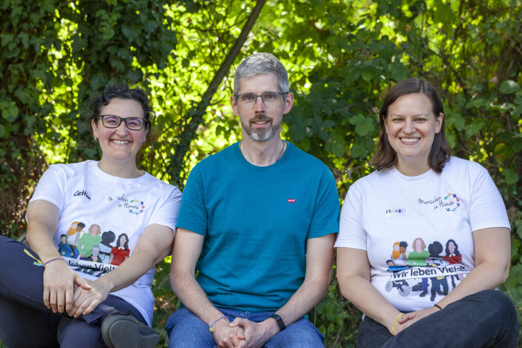 Daniel mit blauem Tshirt und kurzen Haaren sitzt zwischen Cettina und Franzi jeweils mit weißem MIH Tshirt im Grünen.