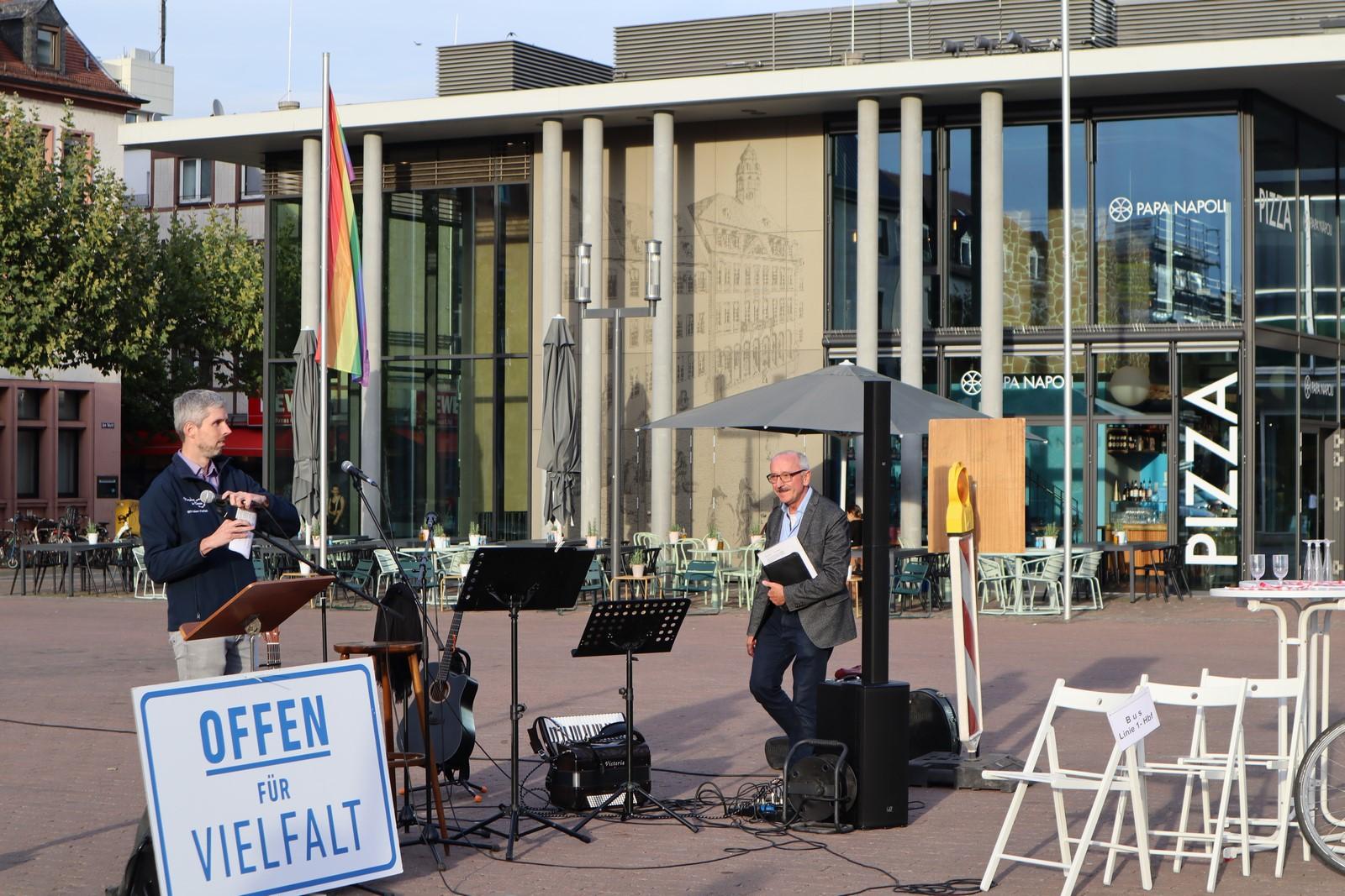 Zu sehen ist Pfarrer Rühl am rechten Ende des Bildes vor der Musikanlage. Daniel nimmt sich das Mikrofon für die Eröffnungsworte. Unten steht auf einem Schild groß "Offen für Vielfalt".