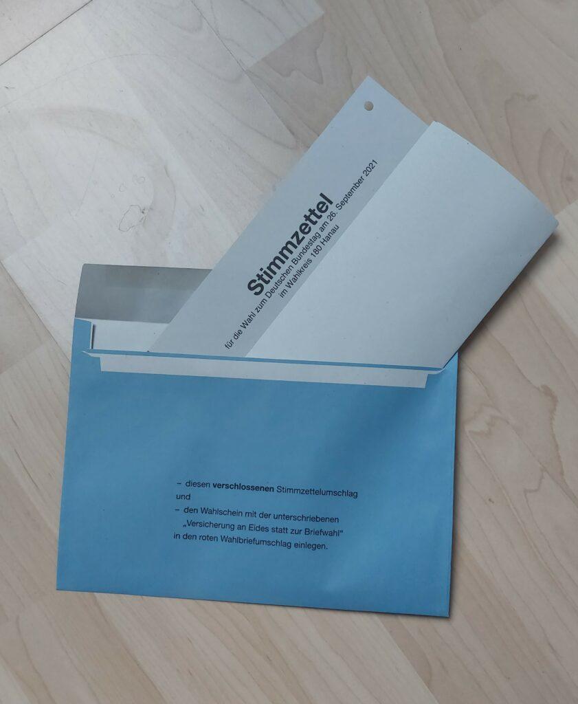 Zu sehen ist der gefaltete Stimmzettel, der in den kleinen blauen Umschlag gesteckt wird.