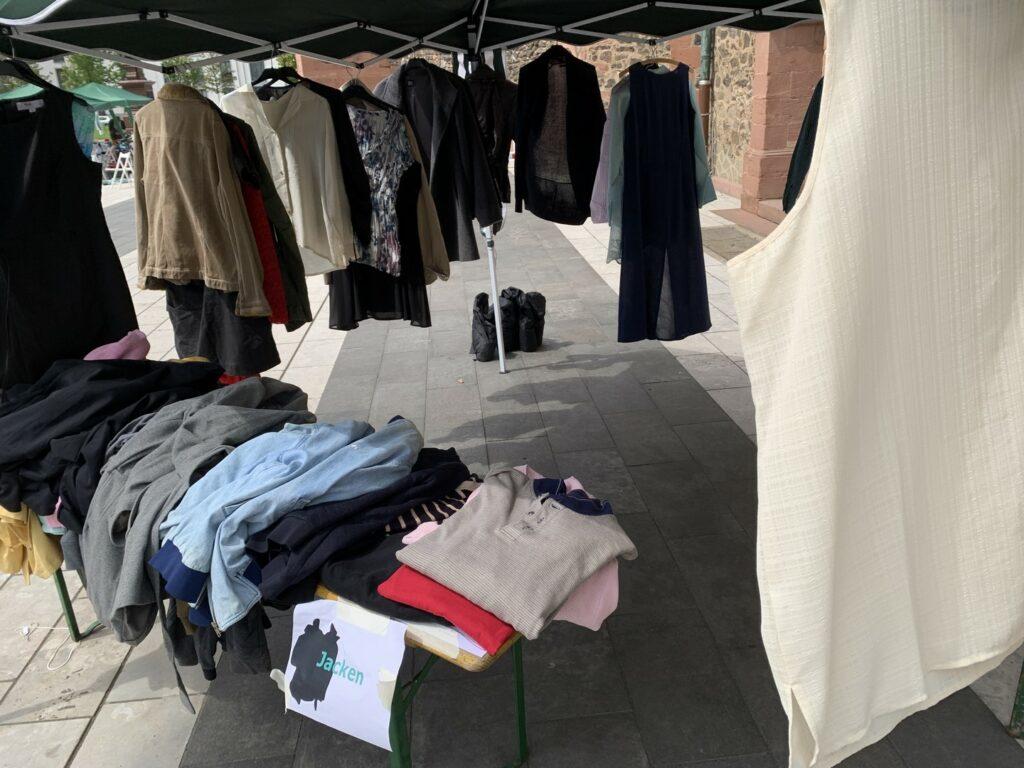 Auf dem Bild sieht man diverse Kleidungsstücke unter einem Zelt. Viele liegen auf einem Tisch, aber Blusen und Jacken hängen auch auf Bügeln an dem Zelt.