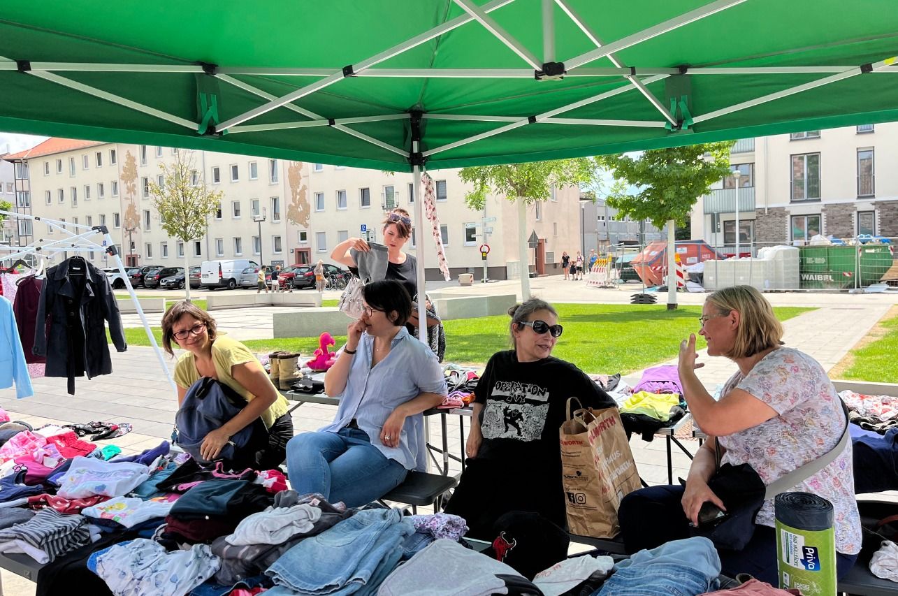 4 Frauen sitzen unter einem Zelt beim Tauschmarkt mit jede Menge Klamotten vor sich und unterhalten sich.