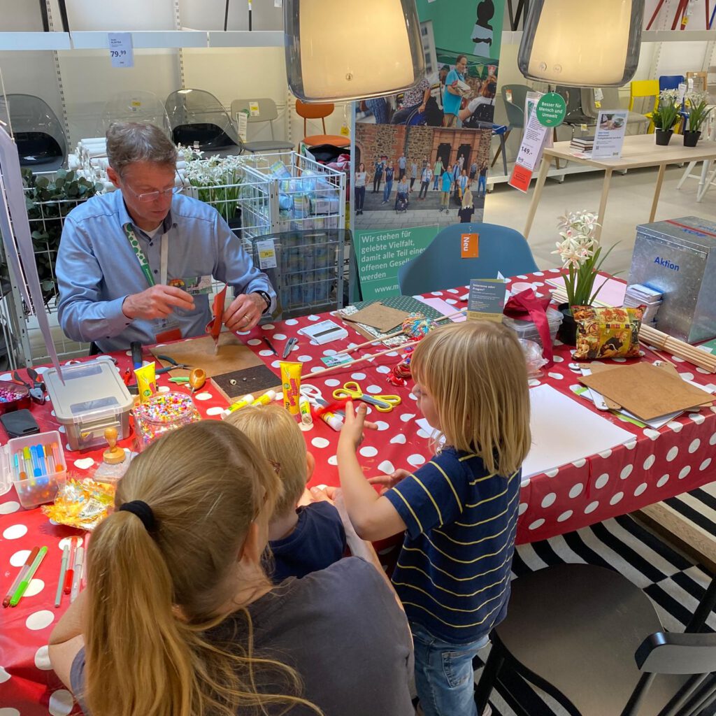 Jochen sitzt an einem Tisch und bastelt. 2 Kinder basteln mit.