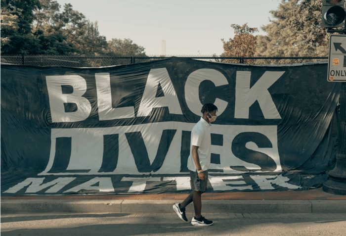 An einem Zaun ist ein großes schwarzes Banner angebracht auf dem mit weißen Buchstaben "Black lives Matter" steht. Ein junger schwarzer Mann läuft im Profil daran vorbei.