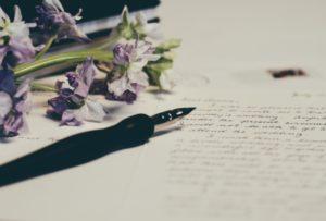Ein handschriftlicher Brief füllt das Bild. Auf dem Papier liegt ein Füller und eine Blume mit lila Blüten.