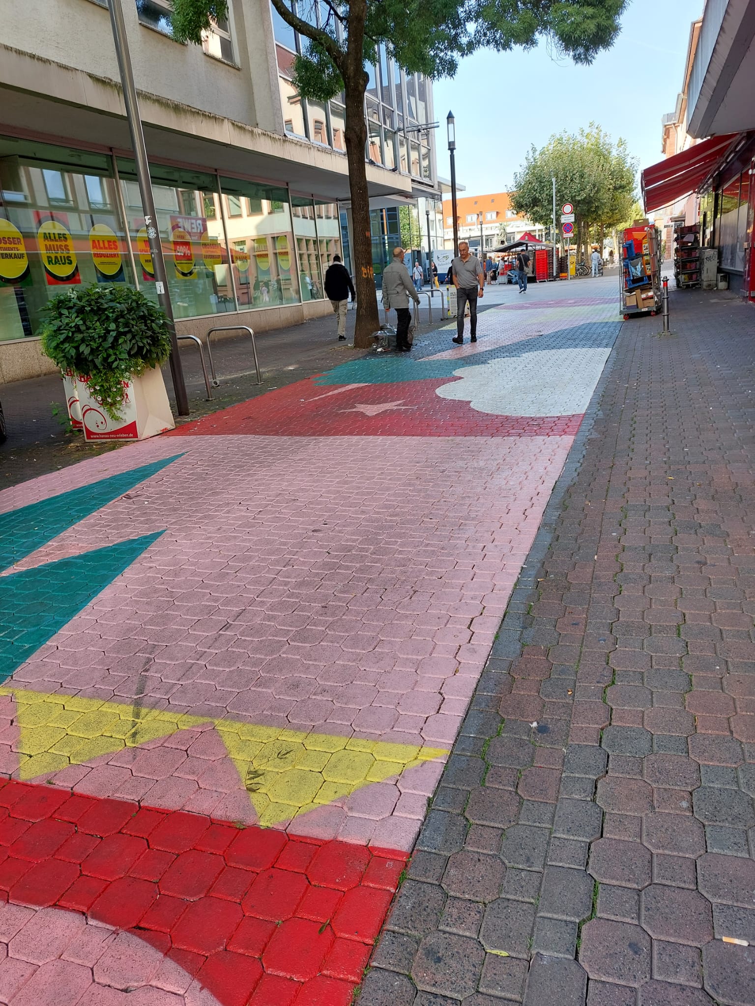Eine Strasse in einer Fussgängerzone ist mit bunten Formen bemalt. Hier sind insbesondere gelbe und grüne Dreeicke zu sehen auf rosafarbenen Hintergrund.