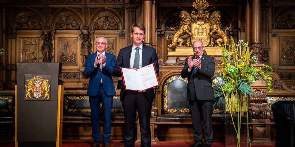 Auf dem Foto ist Botond Roska in einem prunkvollen Raum des Hamburger Rathauses zu sehen. Er steht vorne und präsentiert stolz die Urkunde in rotem Buch. Hinten applaudieren zwei Herren.