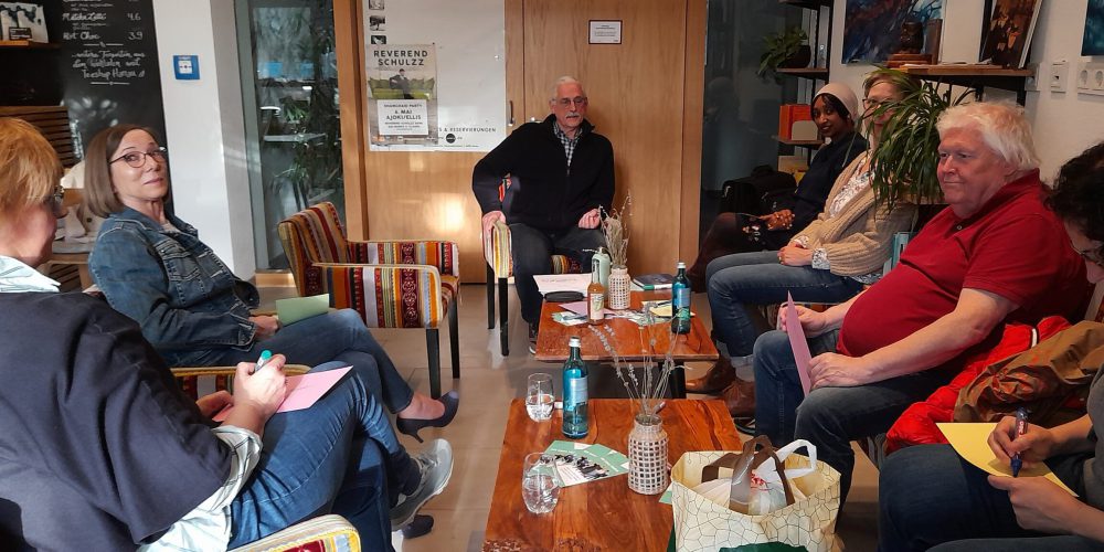 Fünf Frauen und zwei Männer sitzen auf Sessel bzw. auf einem Sofa in einem Café. Sie diskutieren.