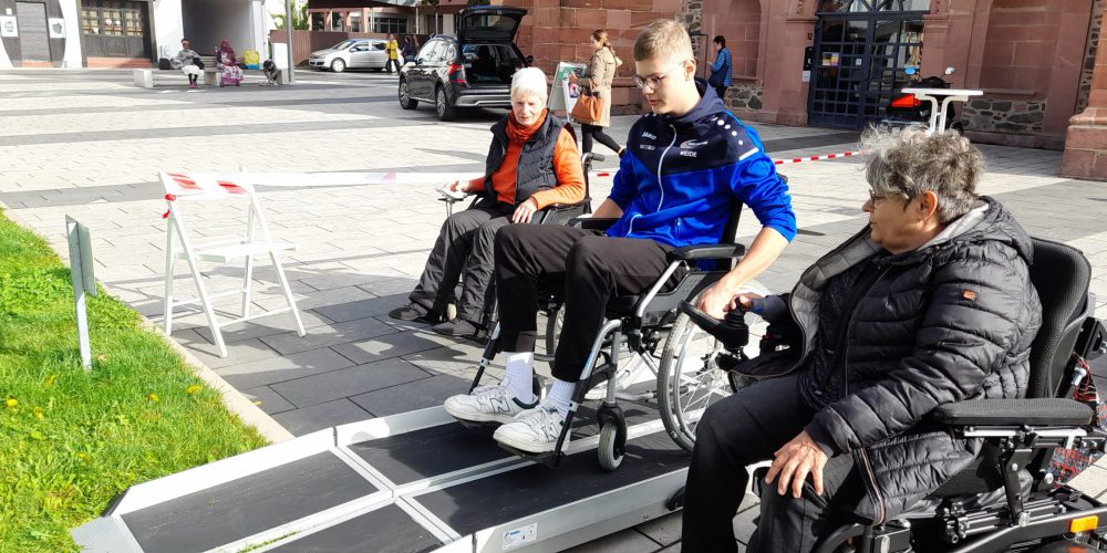 Zwei Frauen und ein Junge, alle im Rollstuhl, sind vor einer Rollstuhlrampe. Die Frauen erklären dem Jugendlichen, wie er die Rampe befahren kann.