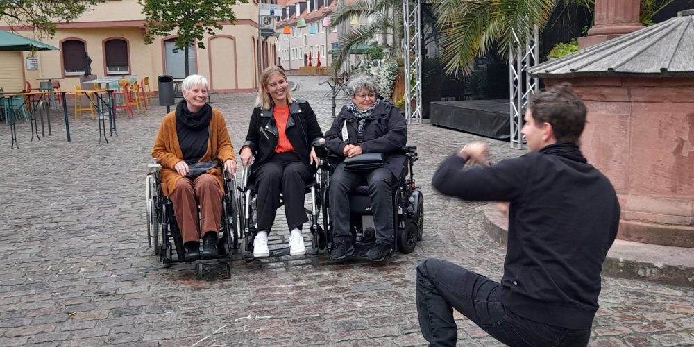 Bärbel, Gaby und die Moderatorin sitzen nebeneinander im Rollstuhl. Ein Mann in schwarzen Klamotten kniet vor ihnen für ein Foto.