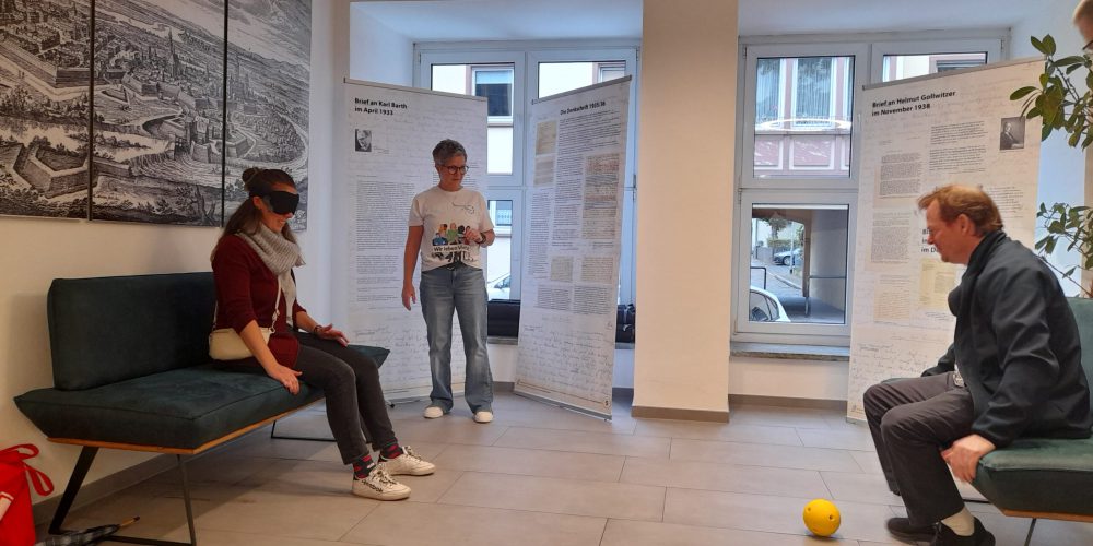 Eine Frau mit Augenbinde sitzt auf einem Sofa. Ihr gegenüber sitzt ein Mann, der sehbehindert ist. In der Mitte steht eine Frau. Vor dem Mann liegt ein gelber Ball. Sie spielen Blindenfussball.