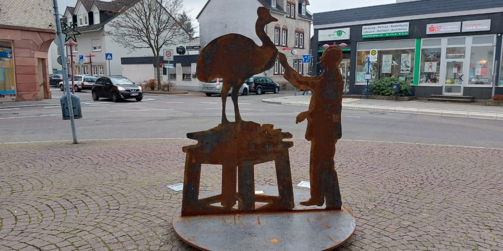 Zu sehen ist ein Mensch der einen auf einem Podest stehenden Vogelstrauus mit einem Werkzeug berabeitet. Die Skulptur ist ein Scherenschnitt und steht auf einer Platte.