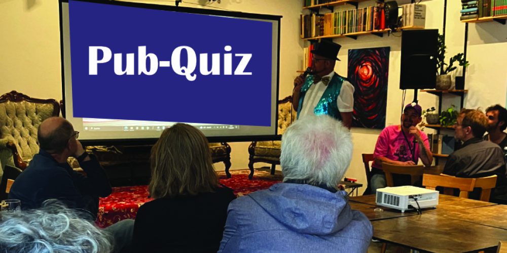 In einem Raum sitzen mehrere Gruppen an Tischen und schauen auf eine Leinwand, auf der Pub-Quiz steht.